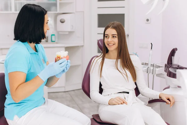女孩坐在牙科椅子上看女牙医 展示了模特的龋状物 — 图库照片