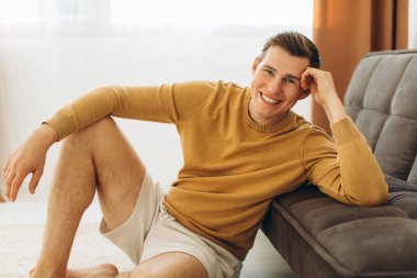 Sarı günlük elbiseli bir adam evde oturuyor ve gülümsüyor.