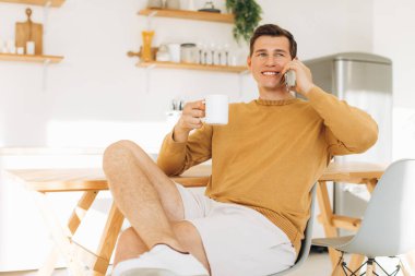 Sarı günlük elbiseli yakışıklı adam mutfakta oturmuş kahve içiyor ve tablet ve telefon üzerinde çalışıyor.