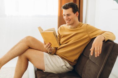 Sarı elbiseli yakışıklı genç adam evde oturmuş kitap okuyor.