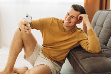 Sarı giysili yakışıklı genç adam evde oturmuş telefonda bir mesaj okuyor.