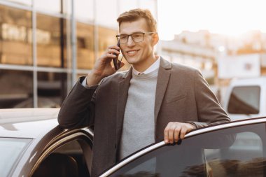 Gözlüklü yakışıklı genç adam telefonla konuşuyor ve dışarıda dikilirken arabasına biniyor.