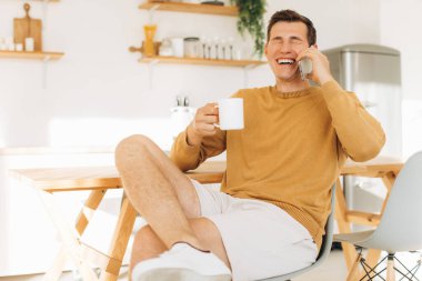Sarı günlük elbiseli yakışıklı adam mutfakta oturmuş kahve içiyor ve tablet ve telefon üzerinde çalışıyor.