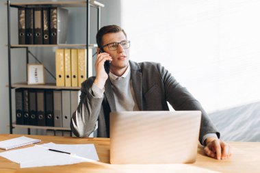 Gözlüklü modern adam gülümsüyor, ofis çalışanı dizüstü bilgisayarda çalışıyor ve telefon görüşmesi yapıyor.