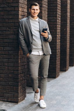 Modern yakışıklı, gülümseyen bir adam. Elinde telefon olan bir duvarın yanında duruyor.