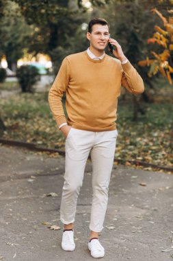 Sarı ve beyaz elbiseli modern yakışıklı adam gün batımında parkta telefonla konuşuyor.