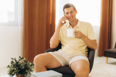 Sarı giysili yakışıklı bir adam evde oturmuş telefonda konuşuyor.