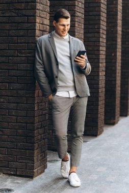 Modern yakışıklı, gülümseyen bir adam. Elinde telefon olan bir duvarın yanında duruyor.