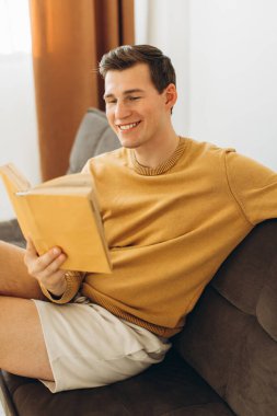 Sarı elbiseli yakışıklı genç adam evde oturmuş kitap okuyor.