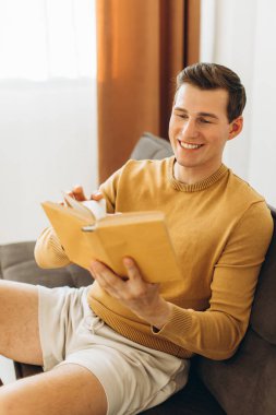 Sarı elbiseli yakışıklı genç adam evde oturmuş kitap okuyor.