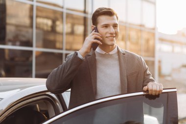 Yakışıklı, modern bir genç adam telefonla konuşuyor ve dışarıda dururken arabasına biniyor.