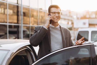 Gözlüklü yakışıklı genç adam telefonla konuşuyor ve dışarıda dikilirken arabasına biniyor.