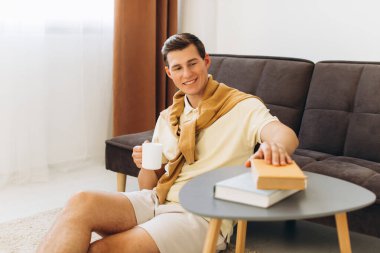 Sarı elbiseli yakışıklı genç adam evde oturmuş kitap okuyor ve kahve içiyor.