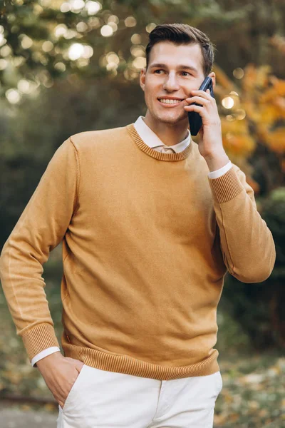 夕暮れ時公園で電話で話している黄色と白の服を着た現代のハンサムな笑顔の男 — ストック写真
