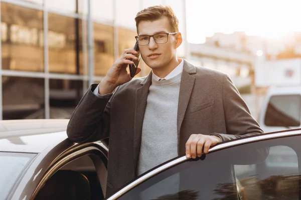 一个戴眼镜的英俊的现代年轻人站在外面 一边打电话一边上车 — 图库照片