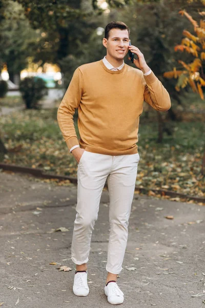 Sarı ve beyaz elbiseli modern yakışıklı adam gün batımında parkta telefonla konuşuyor.