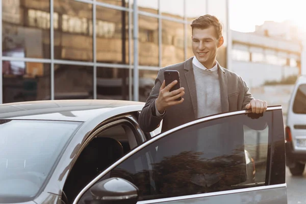 携帯電話を持つハンサムな現代の若者屋外に立っている間に彼の車に入る — ストック写真