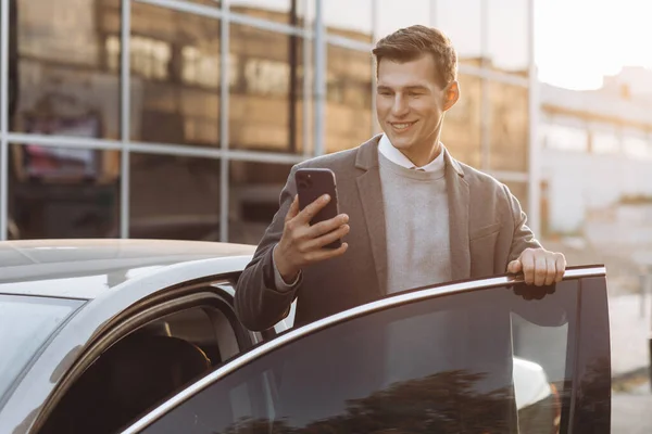 携帯電話を持つハンサムな現代の若者屋外に立っている間に彼の車に入る — ストック写真