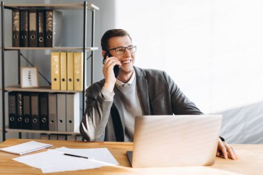 Gözlüklü modern adam gülümsüyor, ofis çalışanı dizüstü bilgisayarda çalışıyor ve telefon görüşmesi yapıyor.