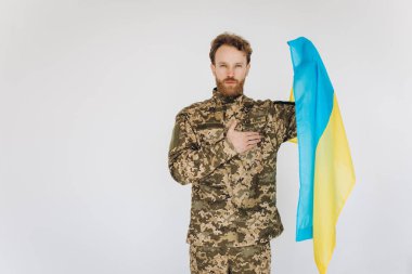 Askeri üniformalı Ukraynalı vatansever asker, beyaz arka planda sarı ve mavi bayrağı olan bir kalbe el kaldırdı.