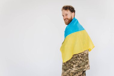 Askeri üniformalı mutlu Ukraynalı asker beyaz arka planda sarı ve mavi bir bayrak tutuyor.