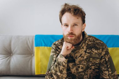 Askeri üniformalı genç bir Ukraynalı vatansever askerin portresi. Üzerinde sarı ve mavi bir bayrak olan bir kanepede oturuyor..