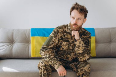 Askeri üniformalı genç bir Ukraynalı vatansever askerin portresi. Üzerinde sarı ve mavi bir bayrak olan bir kanepede oturuyor..