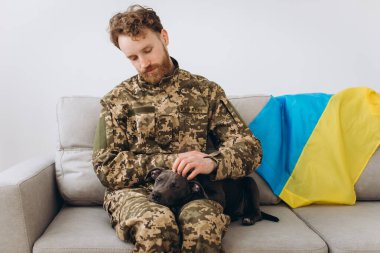 Askeri üniformalı bir Ukraynalı asker sadık arkadaşı Amstaff köpeğiyle birlikte bir kanepede oturuyor. Arka planda sarı ve mavi bayrak..