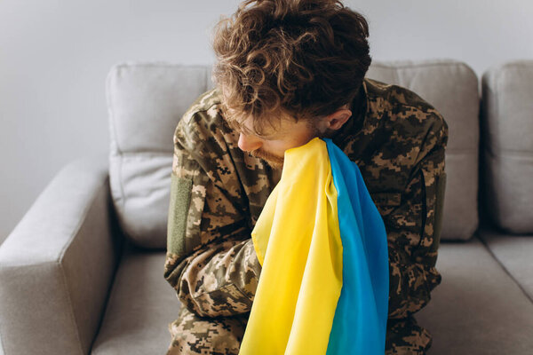 Portrait of an emotional young Ukrainian patriot soldier in military uniform sitting on the office on the couch holding a yellow and blue flag.