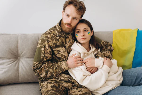 Ukrainian couple, military man in uniform with his girlfriend on the couch at home on a background of yellow and blue flag