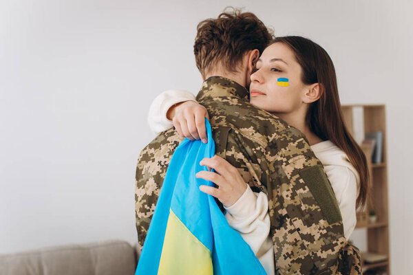 A Ukrainian girl hugs and holds a yellow and blue flag of a military man in uniform and says goodbye to him.