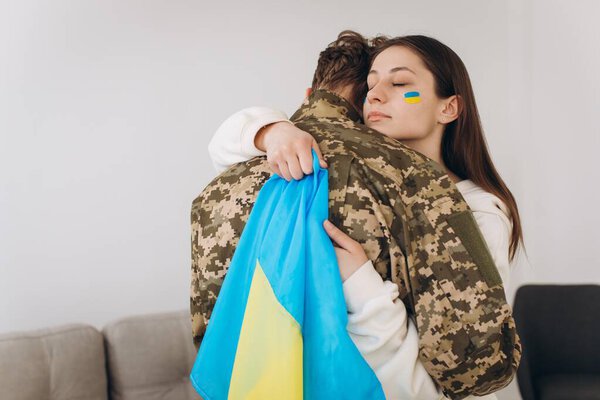 A Ukrainian girl hugs and holds a yellow and blue flag of a military man in uniform and says goodbye to him.