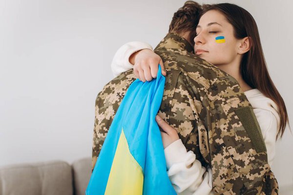A Ukrainian girl hugs and holds a yellow and blue flag of a military man in uniform and says goodbye to him.