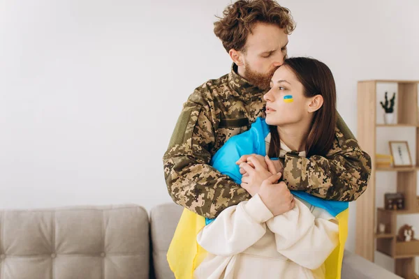 Ukrainian couple, military man in uniform hugs and wraps his wife in the Ukrainian flag. The concept of patriotism.