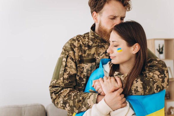 Ukrainian couple, military man in uniform hugs and wraps his wife in the Ukrainian flag. The concept of patriotism.