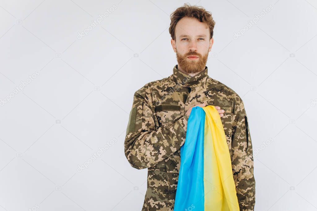 Ukrainian patriot soldier in military uniform holds a hand on a heart with a yellow and blue flag on a white background