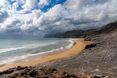 Dağları olan kayalık bir okyanus kıyısı ve ön planda güzel altın bir kumsal.