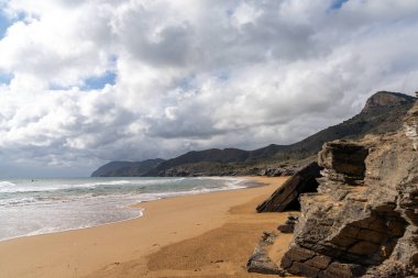 Dağları olan kayalık bir okyanus kıyısı ve ön planda güzel altın bir kumsal.
