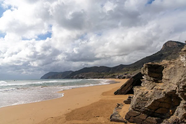 Dağları olan kayalık bir okyanus kıyısı ve ön planda güzel altın bir kumsal.