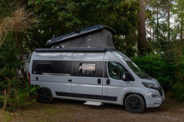 A gray camper van with pop top parked in a sheltered corner of a green forest