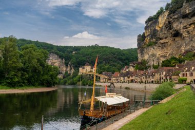 La Roque-Gageac, Fransa - 12 Mayıs 2022: Dordogne Nehri ve uçurum kenarındaki evleri ve ön planda ahşap bir Gabare nehir teknesi olan tarihi La Roque-Gageac köyünün manzarası