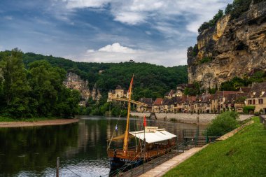 La Roque-Gageac, Fransa - 12 Mayıs 2022: Dordogne Nehri ve uçurum kenarındaki evleri ve ön planda ahşap bir Gabare nehir teknesi olan tarihi La Roque-Gageac köyünün manzarası