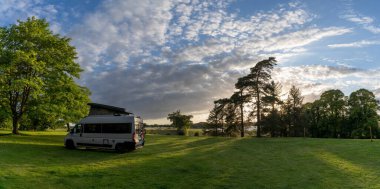 Ormanın manzarası ve akşam ışığında yeşil çayır. Ortasında üstü açılır çatısı olan gri bir karavan.
