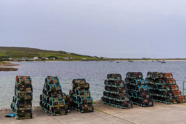 Many stacks of cleaned crab traps and lobster pots on the dock of Blacksod harbor with boats anchored in the bay in the background