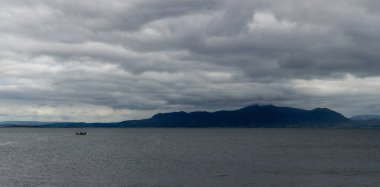 A single fishing boat in a deep blue ocean under a cloudy sky with coastal mountains in the background