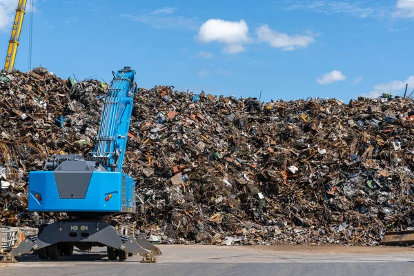 A large pile of scrap metal and waste at a recycling plant with a blue excavator in front