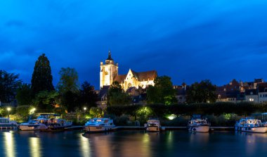 Dole, Fransa - 14 Eylül 2022: Dole 'daki aydınlatılmış Notre Dame Katolik Kilisesi' nin gece görüşü ve ön planda Doubs Nehri 'nde yüzen yüzen yüzen evler.