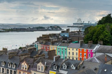 Cobh, İrlanda - 15 Ağustos 2022: Cobh şehir merkezindeki renkli evler Cork Limanı 'na yanaşmış büyük bir yolcu gemisi.