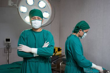 Ameliyat odasındaki doktorlar yeşil maskeli cerrah saçı kep eldiveni giyiyorlar. Biri kolları yatağın önünde çapraz duran, diğeri de elektrikli defibrilatör takıyor. Bu arada doktor da aletleri hazırlıyor.