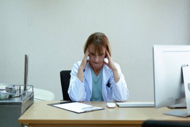 Açık mavi gömlekli, beyaz ceketli, kısa boylu, sarışın kadın doktor. Kablosuz bilgisayar stresinin ve baş ağrısının önünde parlak bir ofiste siyah koltukta oturuyor. Dizüstü bilgisayarın yanında çok zor bir durum var.
